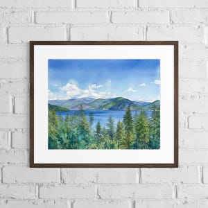 May include: A framed watercolor painting depicting a serene landscape with a deep blue lake, lush green pine trees in the foreground, and rolling mountains under a bright blue sky with scattered white clouds, all enclosed in a dark wood frame.