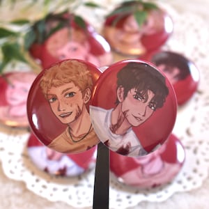 Könnte beinhalten: Eine Sammlung roter und weißer runder Knöpfe mit Illustrationen von zwei jungen Männern mit Verletzungen. Die Knöpfe haben eine glänzende Oberfläche und werden von einem schwarzen Stab gehalten. Weitere Knöpfe mit ähnlichen Designs befinden sich im Hintergrund.