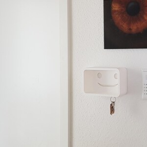 Puede incluir: Un llavero blanco montado en la pared con un diseño de cara sonriente. El llavero tiene forma rectangular y una pequeña balda. Un juego de llaves cuelga de la parte inferior. También se ve un intercomunicador blanco y una obra de arte enmarcada.