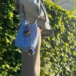 May include: A blue crochet bucket bag with a drawstring closure and a decorative pom-pom. The bag has a long strap and is being carried over the shoulder. The bag is made of a thick, textured yarn.