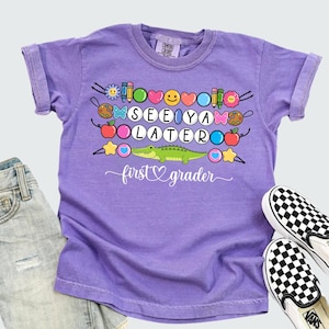 May include: A violet short-sleeved t-shirt with the words "SEE YA LATER first grader" and a cartoon alligator design. The shirt is paired with denim shorts and black and white checkered slip-on shoes.