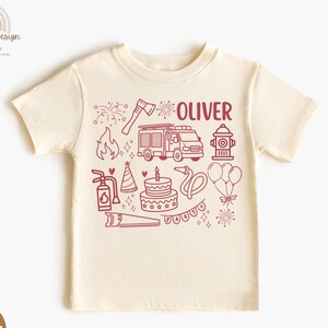 May include: A cream-colored t-shirt featuring a red graphic design with the name "OLIVER". The design includes a fire truck, fire hydrant, cake, and other fire-related illustrations. The shirt is made of a soft material.