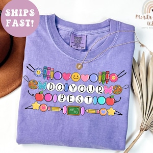May include: A violet-colored t-shirt with the words "DO YOUR BEST" in a playful font, surrounded by colorful school-themed graphics. The shirt has a crew neck and short sleeves. A gold necklace is draped over the shirt. The shirt is labeled "Comfort Colors".