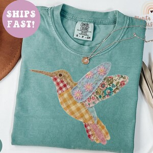 May include: A light green Comfort Colors t-shirt featuring a colorful hummingbird applique. The hummingbird has patchwork wings with floral and gingham patterns. A silver necklace with a small pendant rests on the shirt. The text "SHIPS FAST!" is on a pink circle.