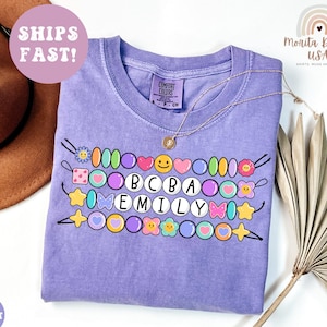 May include: A violet short-sleeved t-shirt featuring a graphic print of three horizontal rows of colourful beads. The beads include shapes like hearts, stars, flowers, and smiley faces. The centre beads spell out the text BCBA and EMILY.
