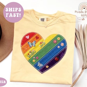 May include: A pale yellow t-shirt featuring a large, central heart-shaped applique. The heart is composed of diagonal rainbow-colored fabric stripes, decorated with embroidered flowers, butterflies, and a ladybug, all outlined with white stitching.