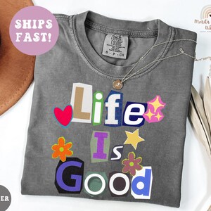 May include: A gray Comfort Colors t-shirt with the phrase "Life Is Good" in a colorful, cut-out style. The design includes a red heart, stars, and flowers. A silver necklace with a pendant is on the shirt.