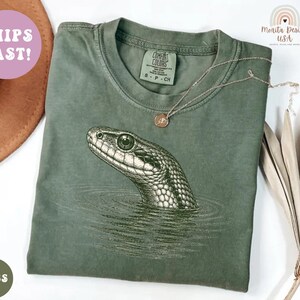 May include: A moss green short-sleeved t-shirt featuring a detailed graphic print of a snake head emerging from water. The shirt has a crew neckline and a label at the inner collar. The design is rendered in a dark ink color, showing the snake's scales and eye clearly.