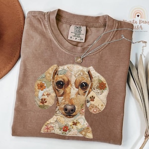 May include: A brown Comfort Colors t-shirt featuring a patchwork dachshund dog design. The dog's ears and face are made of various fabric patterns with floral and paw print accents. A silver necklace with a pendant is draped across the shirt.