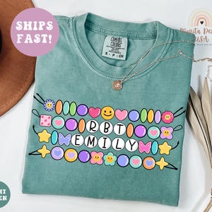 May include: Light green Comfort Colors t-shirt with a beaded design featuring the name "EMILY". The design includes colorful beads, hearts, stars, and flowers. The shirt also has the text "SHIPS FAST!"