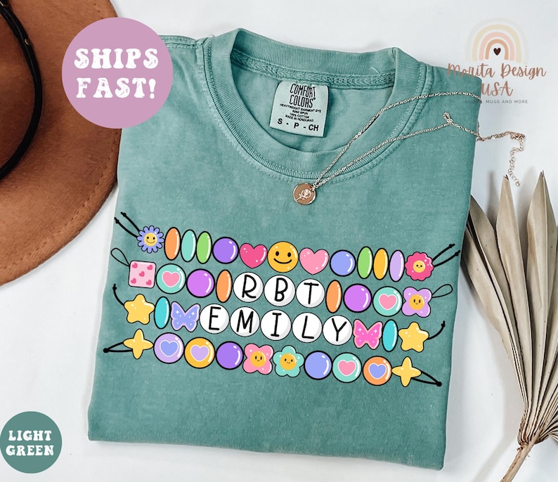May include: Light green Comfort Colors t-shirt with a beaded design featuring the name "EMILY". The design includes colorful beads, hearts, stars, and flowers. The shirt also has the text "SHIPS FAST!"