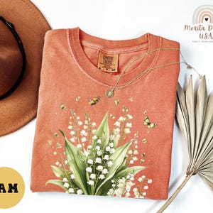 May include: Coral-colored t-shirt featuring a floral design of lily of the valley and butterflies. The shirt has a crew neck and short sleeves. A gold necklace is draped across the shirt. The shirt is from Comfort Colors.