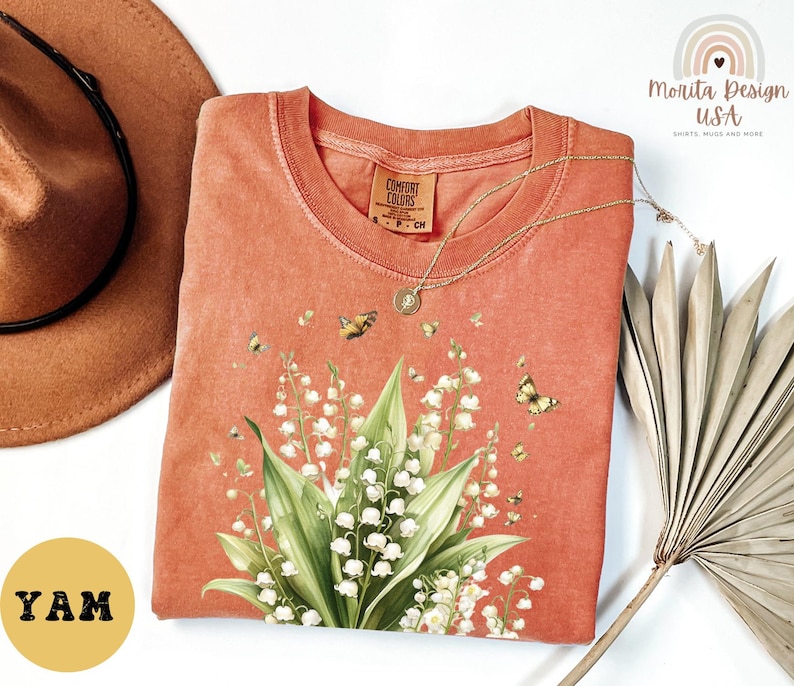 May include: Coral-colored t-shirt featuring a floral design of lily of the valley and butterflies. The shirt has a crew neck and short sleeves. A gold necklace is draped across the shirt. The shirt is from Comfort Colors.