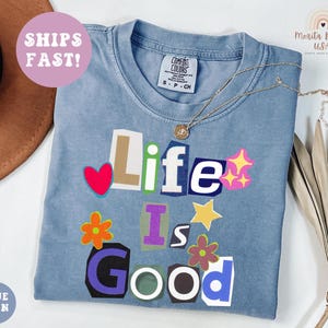 May include: A blue jean colored t-shirt with the words "Life Is Good" in a collage style design. The shirt has a crew neck and short sleeves. A gold necklace with a pendant is draped on the shirt.