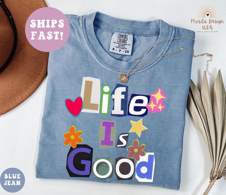 May include: A blue jean colored t-shirt with the words "Life Is Good" in a collage style design. The shirt has a crew neck and short sleeves. A gold necklace with a pendant is draped on the shirt.