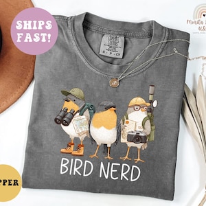 Op de afbeelding: Een grijze t-shirt met een afbeelding van drie cartoonvogels, elk uitgerust met een verrekijker, camera's en kaarten. De tekst "BIRD NERD" staat eronder. De t-shirt heeft een ronde hals en korte mouwen.