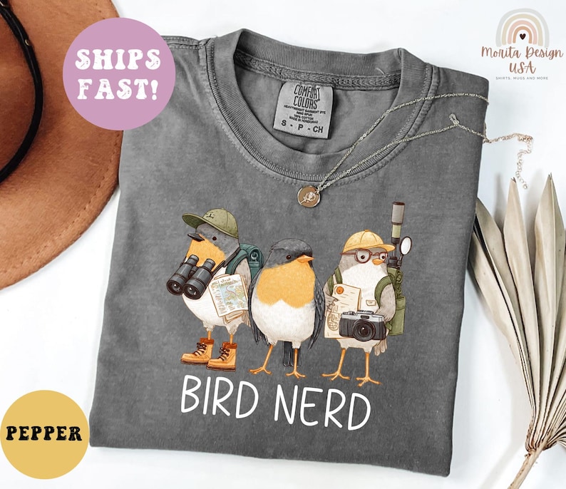 Op de afbeelding: Een grijze t-shirt met een afbeelding van drie cartoonvogels, elk uitgerust met een verrekijker, camera's en kaarten. De tekst "BIRD NERD" staat eronder. De t-shirt heeft een ronde hals en korte mouwen.