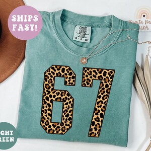 May include: Light green t-shirt with the number 67 in a leopard print design. The shirt has a crew neck and short sleeves. The text "SHIPS FAST!" is on a pink circle. The text "LIGHT GREEN" is on a green circle.