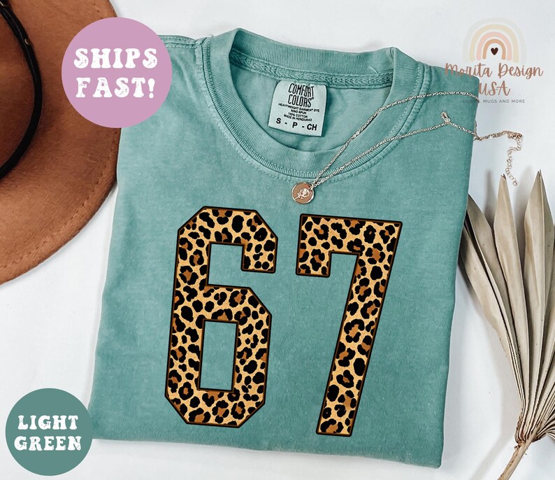 May include: Light green t-shirt with the number 67 in a leopard print design. The shirt has a crew neck and short sleeves. The text "SHIPS FAST!" is on a pink circle. The text "LIGHT GREEN" is on a green circle.