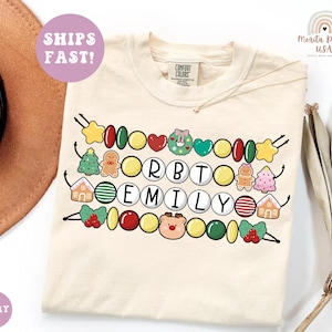May include: An ivory t-shirt featuring a graphic of three horizontal strings of colorful Christmas-themed beads and charms. The beads spell out the name ORBTO EMILY, interspersed with icons of gingerbread people, houses, a wreath, and a reindeer.