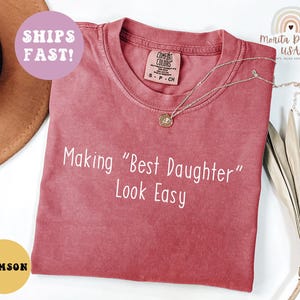 May include: Crimson-colored t-shirt with the text "Making 'Best Daughter' Look Easy" in white. The shirt has a round neckline and short sleeves. A silver necklace with a pendant is on the shirt.