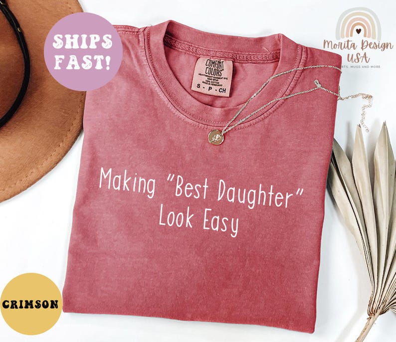 May include: Crimson-colored t-shirt with the text "Making 'Best Daughter' Look Easy" in white. The shirt has a round neckline and short sleeves. A silver necklace with a pendant is on the shirt.