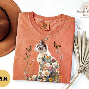 May include: A peach-coloured t-shirt featuring a floral cat design. The cat is surrounded by butterflies and flowers. The shirt has the text "Comfort Colors" on the tag.