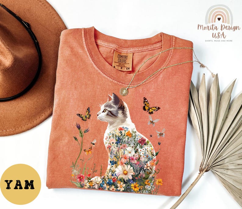 May include: A peach-coloured t-shirt featuring a floral cat design. The cat is surrounded by butterflies and flowers. The shirt has the text "Comfort Colors" on the tag.