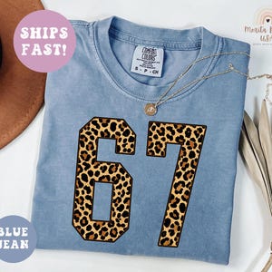 May include: A blue jean colored t-shirt with the number 67 in a leopard print design. The shirt has a crew neck and short sleeves. The text "SHIPS FAST!" is in a pink circle, and "BLUE JEAN" is in a blue circle.