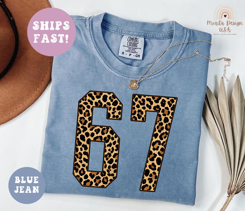 May include: A blue jean colored t-shirt with the number 67 in a leopard print design. The shirt has a crew neck and short sleeves. The text "SHIPS FAST!" is in a pink circle, and "BLUE JEAN" is in a blue circle.