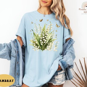 May include: Light blue t-shirt featuring a floral design of lily of the valley and butterflies. The graphic is centered on the shirt and includes green leaves and white flowers. The shirt is made of a soft material.