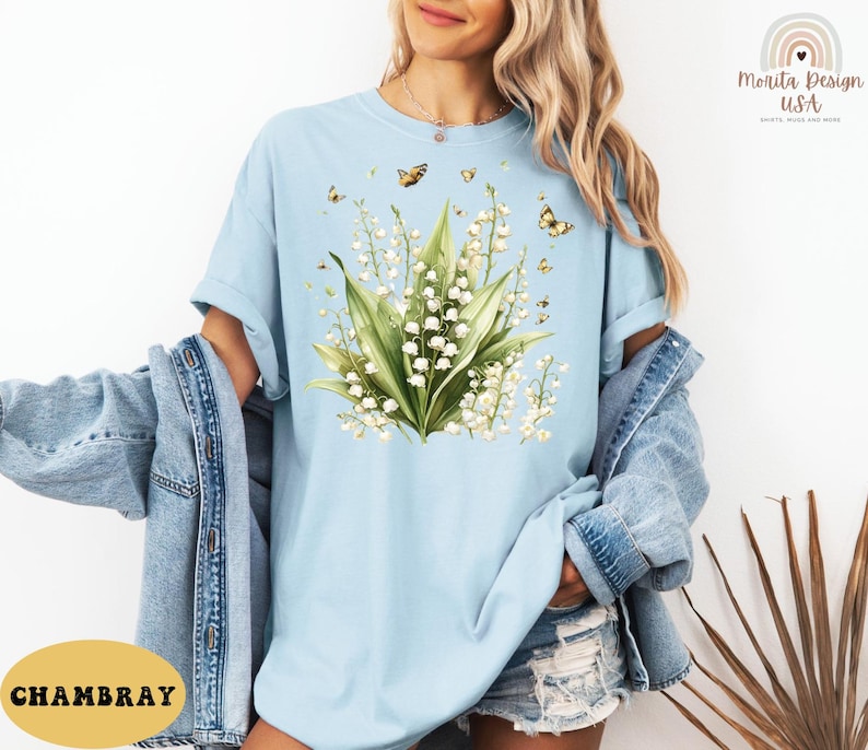 May include: Light blue t-shirt featuring a floral design of lily of the valley and butterflies. The graphic is centered on the shirt and includes green leaves and white flowers. The shirt is made of a soft material.