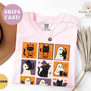 May include: A light pink Comfort Colors t-shirt featuring a Halloween-themed graphic. The graphic includes black cats, ghosts, pumpkins, and bats in orange and purple postage stamp designs. A gold necklace is visible. The shirt is labeled "Blossom."