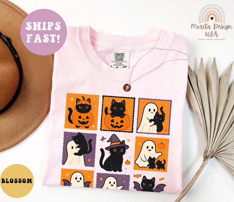 May include: A light pink Comfort Colors t-shirt featuring a Halloween-themed graphic. The graphic includes black cats, ghosts, pumpkins, and bats in orange and purple postage stamp designs. A gold necklace is visible. The shirt is labeled "Blossom."