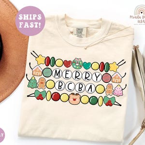 May include: An ivory t-shirt featuring a graphic print of three horizontal strings of colorful beads and holiday-themed charms. The center beads spell out the text MERRY BCBA in white circles. The design includes gingerbread men, houses, and trees.