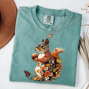 May include: A teal blue t-shirt with a graphic of a squirrel surrounded by flowers. The squirrel is holding a nut in its paws. The t-shirt has a tag that says "Comfort Colors" and "S - P - CH".