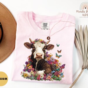 以下が含まれることがあります： ピンクのTシャツに、花冠をかぶった茶色と白の牛が描かれています。牛はカラフルな花と蝶に囲まれています。Tシャツの左下には「Blossom」の文字があります。