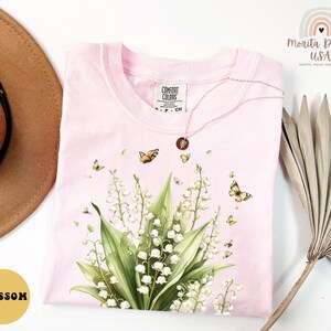 May include: A light pink Comfort Colors t-shirt featuring a floral design with butterflies. The design includes white flowers, green leaves, and several yellow butterflies. A gold necklace with a pendant rests on the shirt. The word "Blossom" is in a yellow circle.