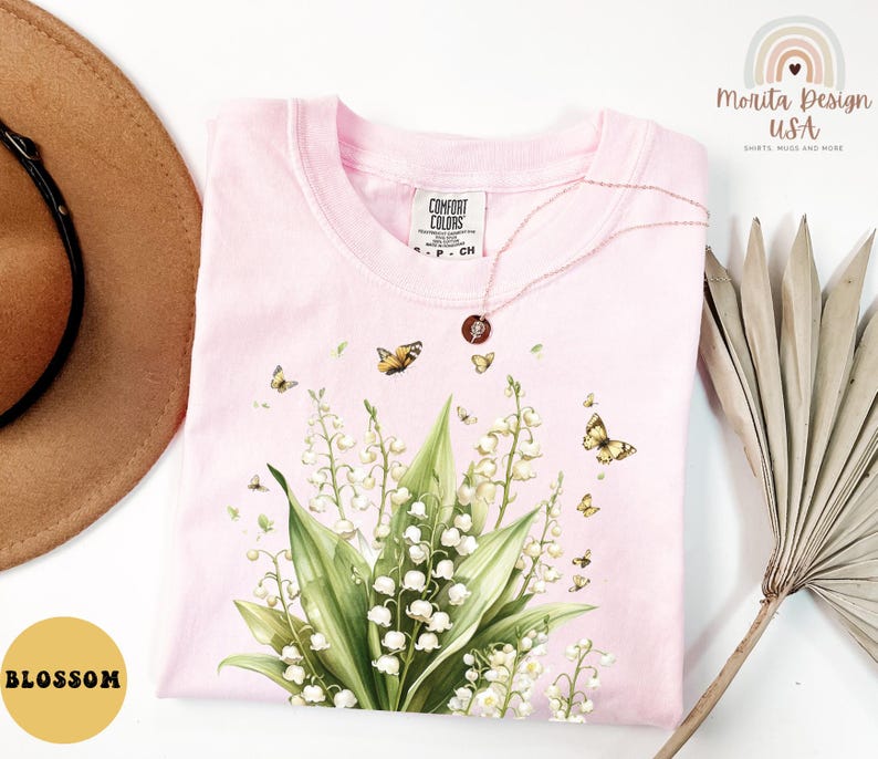May include: A light pink Comfort Colors t-shirt featuring a floral design with butterflies. The design includes white flowers, green leaves, and several yellow butterflies. A gold necklace with a pendant rests on the shirt. The word "Blossom" is in a yellow circle.