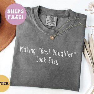 May include: A gray Comfort Colors t-shirt with the text "Making "Best Daughter" Look Easy" in white. A gold necklace is draped across the shirt. A round sticker that says "Pepper" is in the bottom left corner.