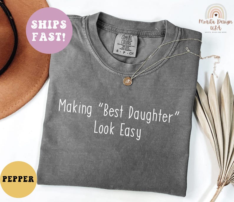 May include: A gray Comfort Colors t-shirt with the text "Making "Best Daughter" Look Easy" in white. A gold necklace is draped across the shirt. A round sticker that says "Pepper" is in the bottom left corner.