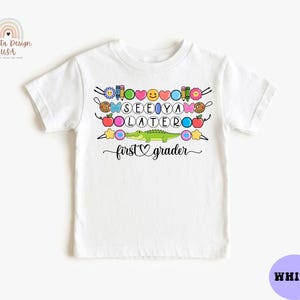 May include: White t-shirt with a colorful graphic that says "SEE YA LATER" with a crocodile and the words "first grader". The design includes various school-themed elements like pencils, apples, and stars.