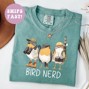 May include: Teal t-shirt with a graphic of three cartoon birds, each with binoculars, cameras, and backpacks. The white text "BIRD NERD" is below the birds. A necklace is draped across the shirt. Measurements are in cm.