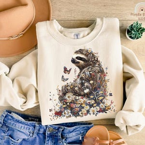 May include: A beige sweatshirt with a graphic print of a sloth surrounded by flowers and butterflies. The sloth is brown and has a floral pattern on its fur. The flowers are in shades of pink, purple, yellow, and white.