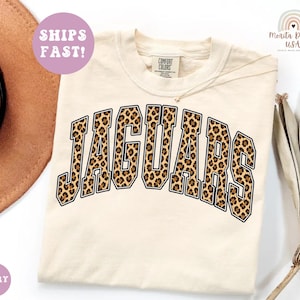 May include: An ivory short-sleeved t-shirt featuring the word JAGUARS in a large, arched, block-letter font with a brown and black leopard print pattern. The shirt has a Comfort Colors brand label at the inner neckline.