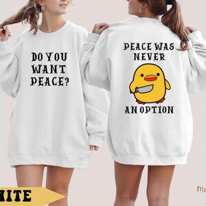 K&ouml;nnte beinhalten: Wei&szlig;er Pullover mit dem Text "Do you want peace?" auf der Vorderseite und "Peace was never an option" mit einem Cartoon-gelben K&uuml;ken, das ein Messer h&auml;lt, auf der R&uuml;ckseite.