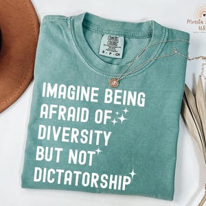 May include: A teal Comfort Colors t-shirt with the text "IMAGINE BEING AFRAID OF DIVERSITY BUT NOT DICTATORSHIP" in white. A gold necklace with a pendant is on the shirt. The shirt is folded on a white surface.