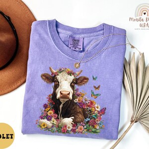 以下が含まれることがあります： 花冠をかぶった牛のフローラルプリントが施されたパープルのTシャツ。牛は蝶と花に囲まれています。シャツは柔らかく快適な素材で作られています。
