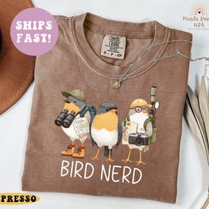 Op de afbeelding: Bruin t-shirt met een afbeelding van drie vogelpersonages met hoeden en uitrusting. De tekst "BIRD NERD" is wit gedrukt. Het shirt heeft een Comfort Colors-label.