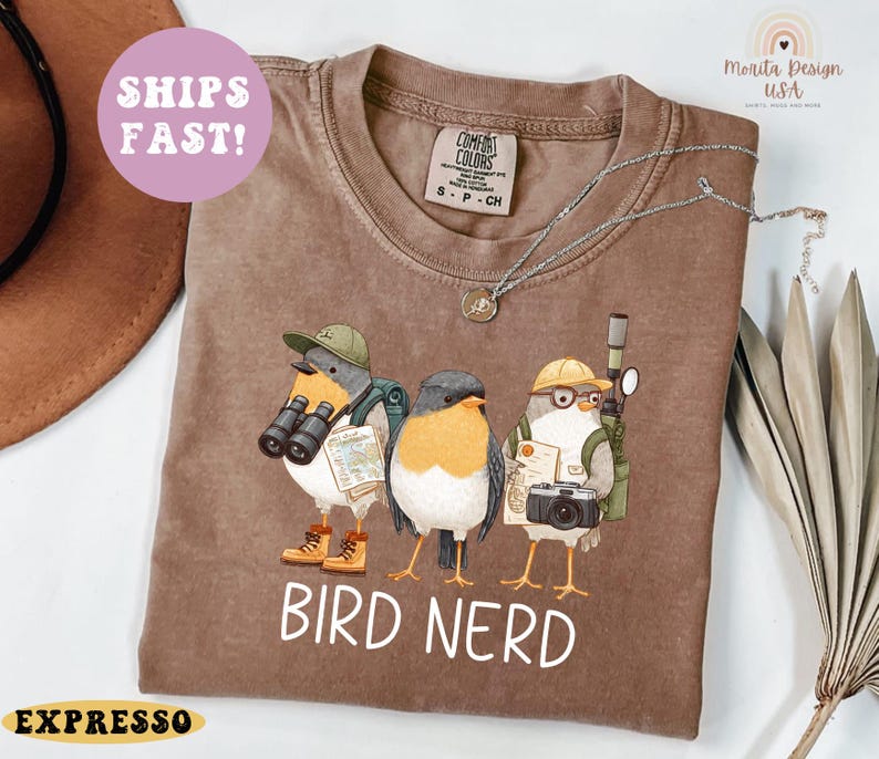 Op de afbeelding: Bruin t-shirt met een afbeelding van drie vogelpersonages met hoeden en uitrusting. De tekst "BIRD NERD" is wit gedrukt. Het shirt heeft een Comfort Colors-label.